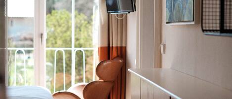 Habitación de hotel con cama, televisor en la pared, una silla y ventana con vistas a zonas verdes.