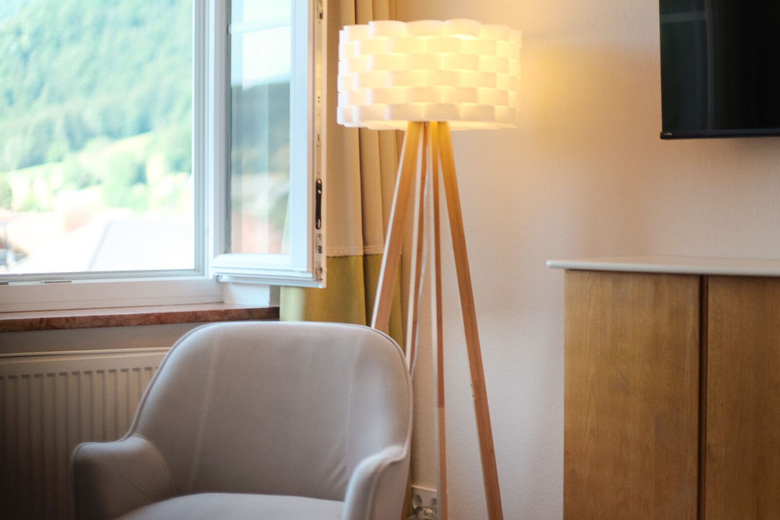A room with a white chair, a floor lamp with a checkered shade, a window with a view of greenery, and a mounted TV on the wall.