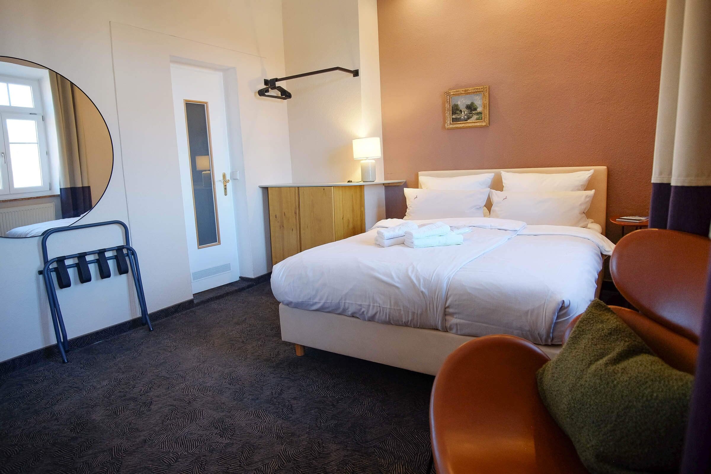 A hotel room with a bed, a chair, a bedside table with a lamp, a mirror, and a door.