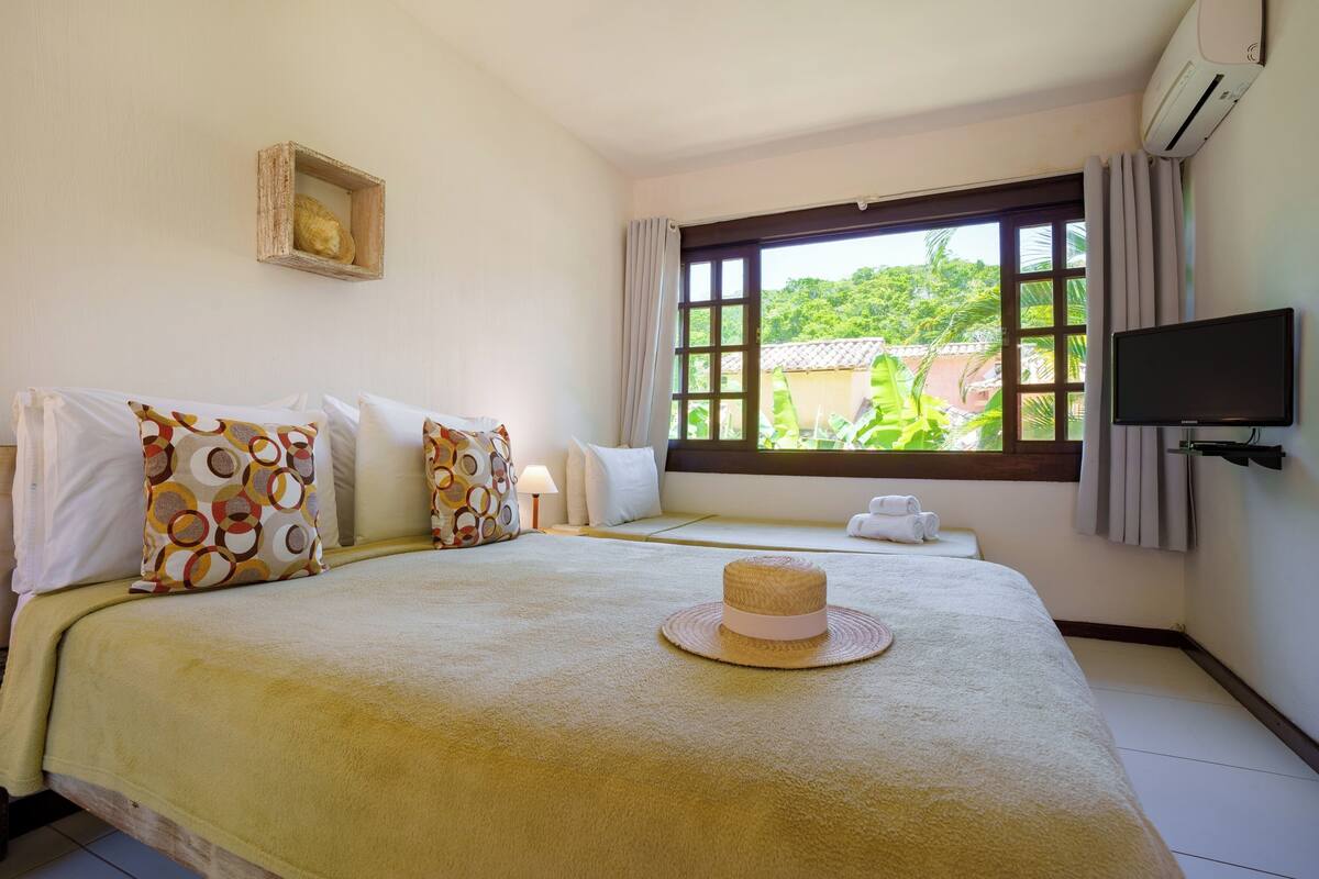 A hotel room with a large bed, a TV, a window with curtains, and a view of greenery outside.