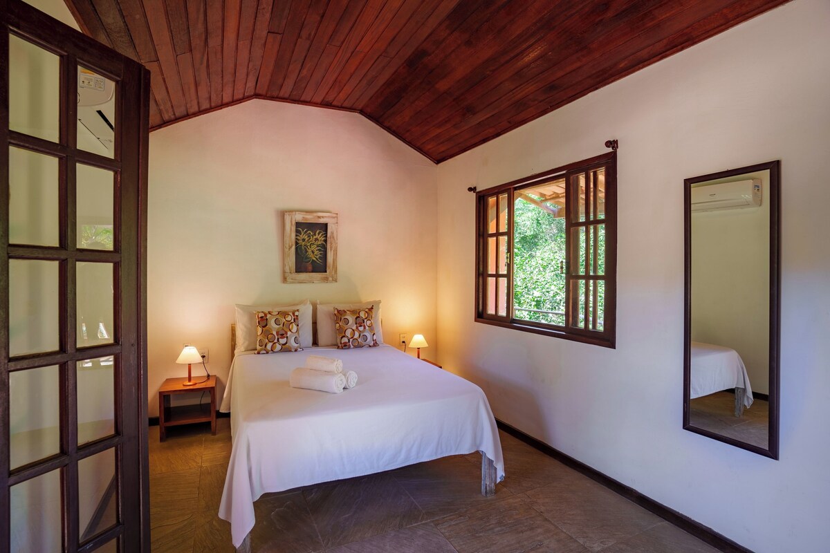 A bedroom with a wooden ceiling, a bed with white bedding, a nightstand with a lamp, a window with a view of greenery, and a wall-mounted air conditioning unit.