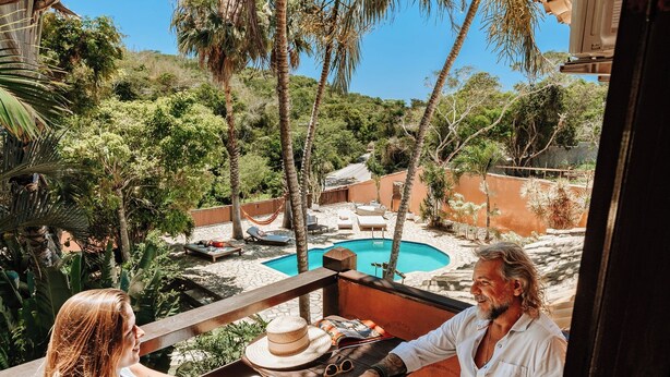 A couple seated at a wooden table on a balcony overlooking a swimming pool, with palm trees and a clear sky in the background. - Pousada Casa Cactus (Búzios)
