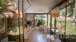 A spacious, sunlit room with a long wooden bench, a small kitchen area, and large windows offering a view of lush greenery. - Pousada Casa Cactus (Búzios)