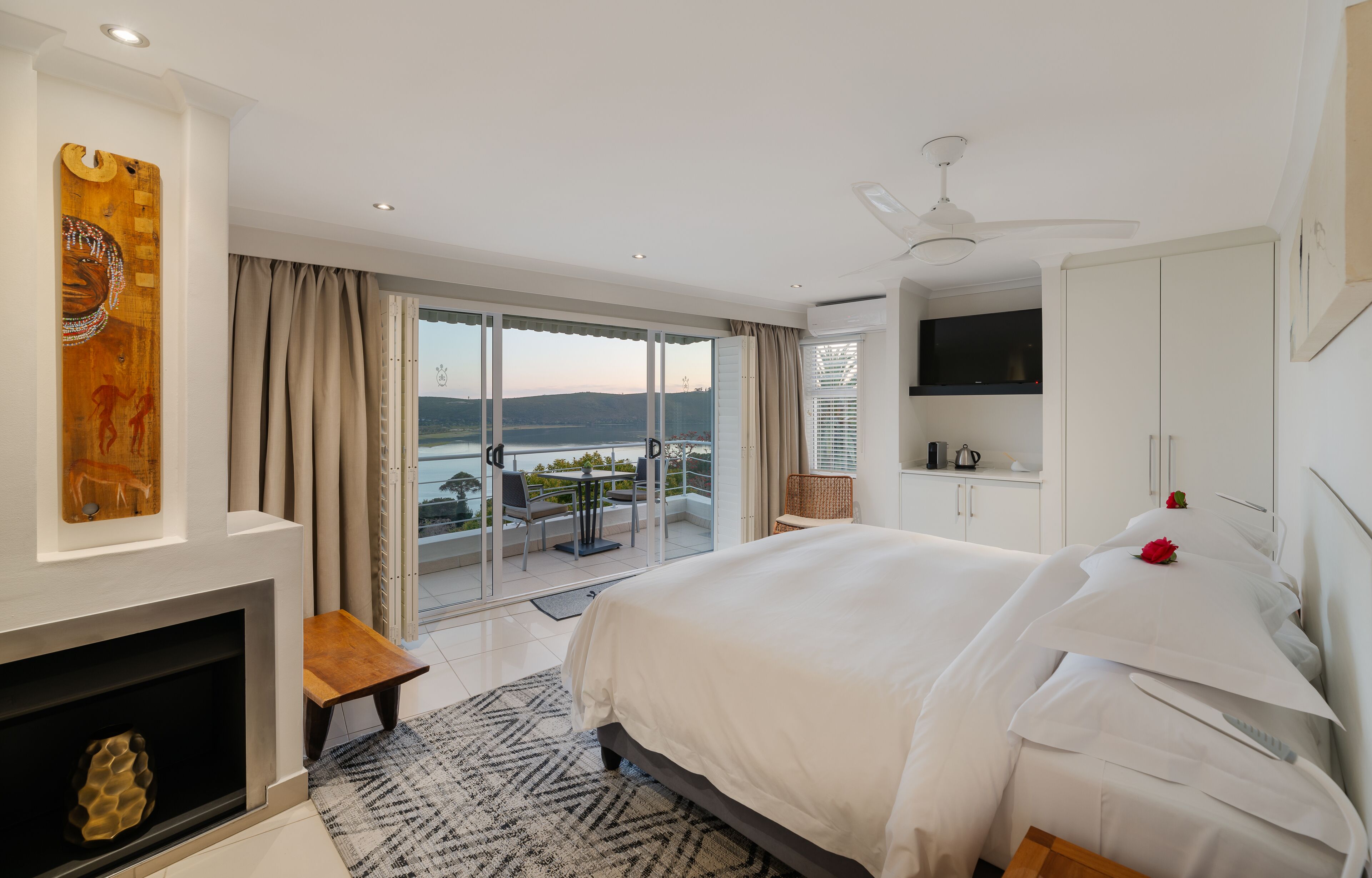 Quarto com cama grande, lareira, vista para o mar e varanda com mesa e cadeiras.