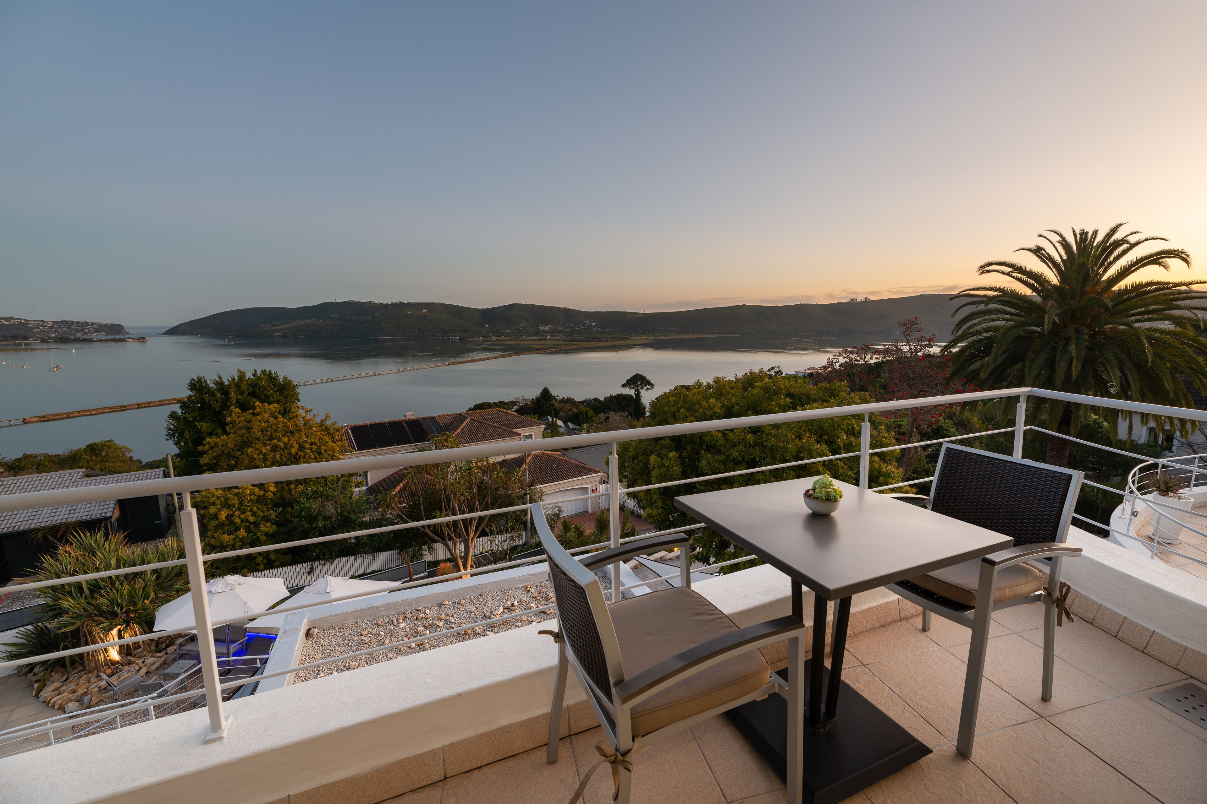 Un balcon avec une table et des chaises donnant sur une étendue d’eau et sur des collines lointaines.