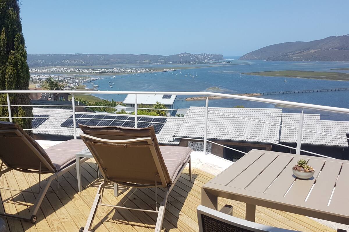 Une terrasse dotée de deux chaises longues et d’une table, offrant une vue sur une marina avec des bateaux et sur les contours d’une ville au loin.