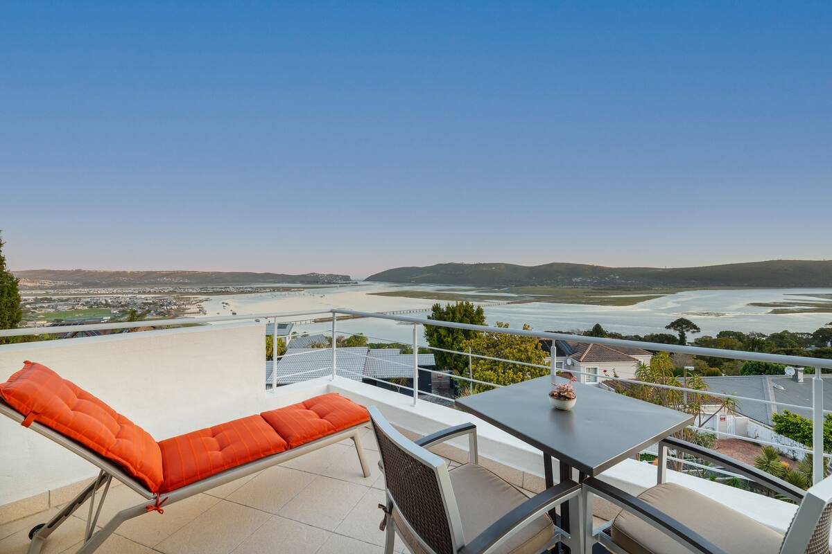 Un balcon donnant sur une rivière et des collines lointaines, une table et un fauteuil avec un coussin orange.