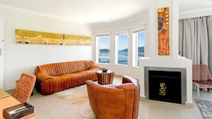 Un salon moderne avec un canapé, un fauteuil et une cheminée. De grandes fenêtres offrent une vue sur la mer.