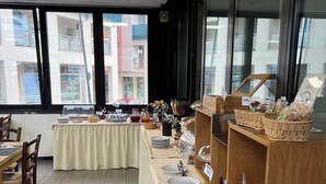 Een buffet met diverse gerechten en drankjes, een eetgedeelte met stoelen en een tafel, en grote ramen met uitzicht op de stad.