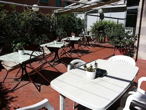 Un espace extérieur aménagé en terrasse, avec des tables et des chaises, entouré de plantes en pot et avec un bâtiment en arrière-plan.