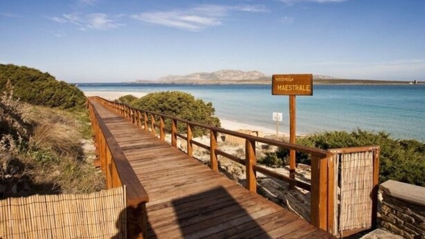 A wooden boardwalk leading to a beach with a sign reading "PAGERELLA MAESTRALE". - Hotel Cala Reale (Stintino)