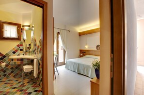 A hotel room with a bathroom featuring colorful tiles, a bed with a light blue bedspread, a bedside table with a plant, and a wooden headboard. - Hotel Cala Reale (Stintino)