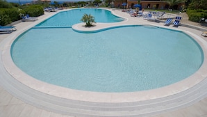 A large, clear swimming pool with blue lounge chairs and umbrellas, surrounded by greenery and buildings. - Hotel Cala Reale (Stintino)
