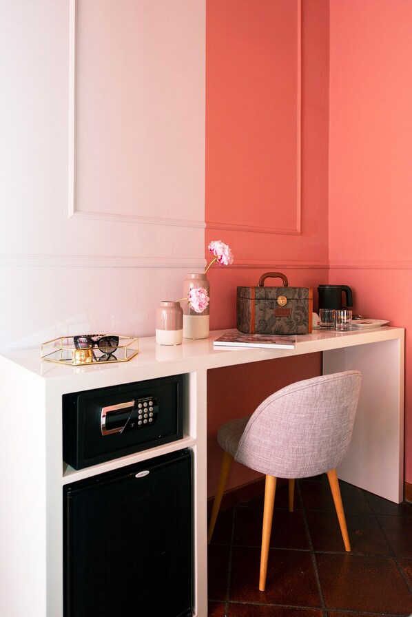 Un espace de travail compact avec un bureau blanc, un mur rose, une chaise grise, un micro-ondes noir, un petit réfrigérateur noir, un vase décoratif avec des fleurs roses et un sac à main à motifs.