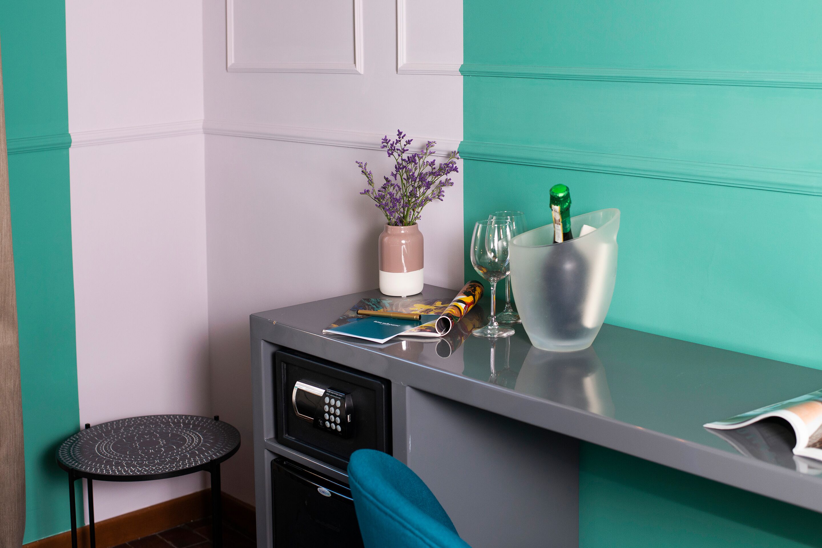 A compact room with teal and white walls, a small table with a vase of flowers, a bottle of champagne, and a few glasses.