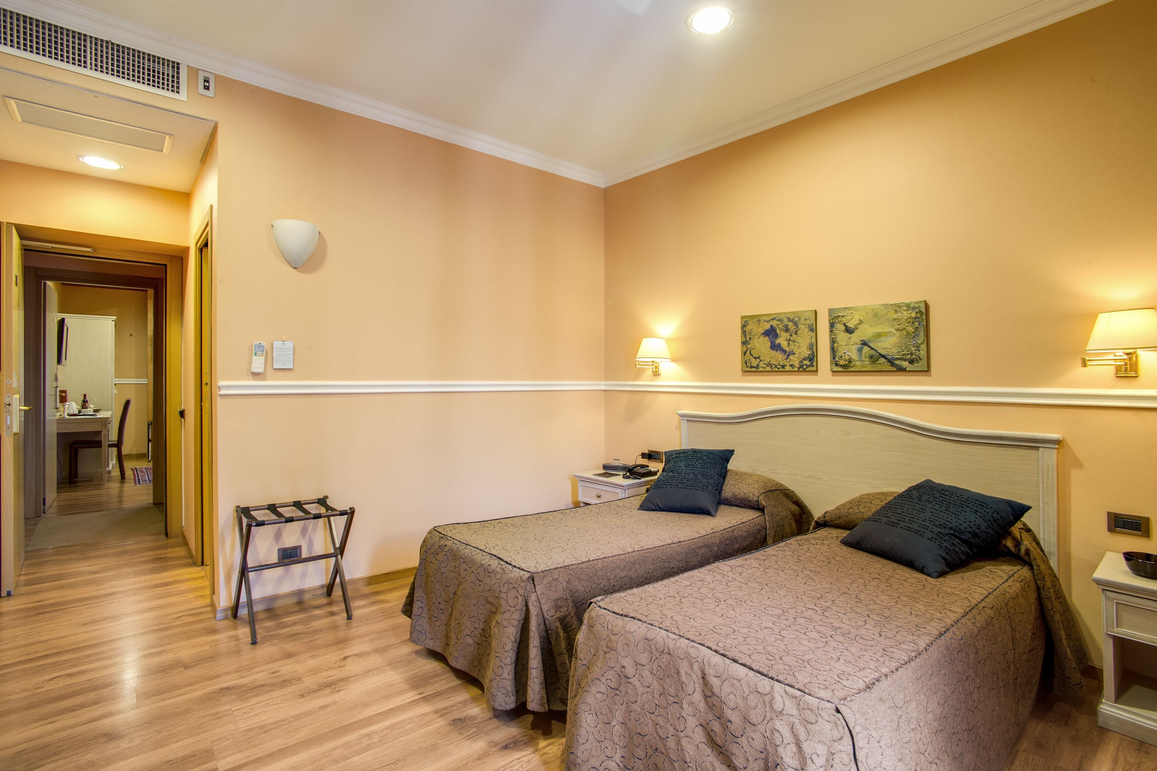 Quarto de hotel com duas camas de solteiro, uma pequena mesa, uma cadeira, um criado-mudo e uma luminária na parede.