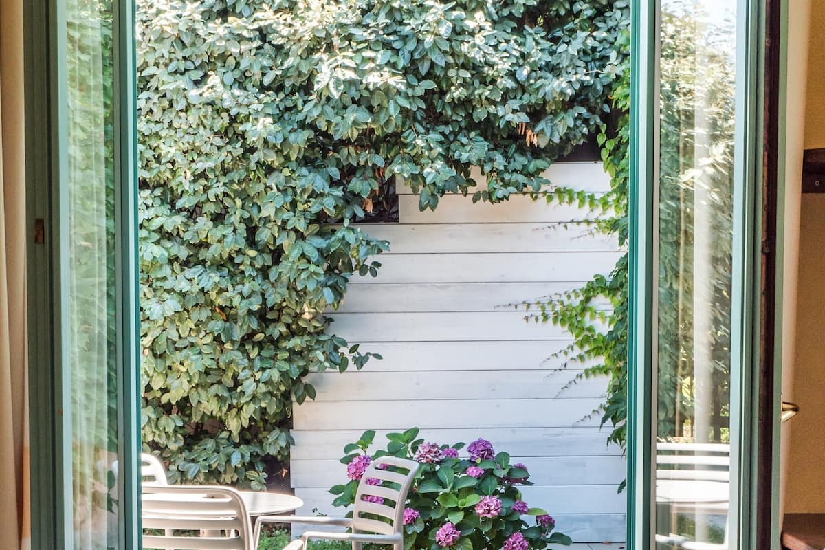 Una puerta corredera de vidrio abierta que da a un patio con un muro blanco de madera, un pequeño jardín con flores rosas y una mesa con sillas.