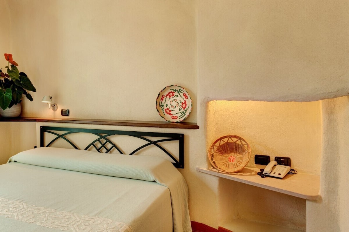 Un dormitorio con una cama, un estante con una cesta y un teléfono, una planta en maceta y un plato decorativo.