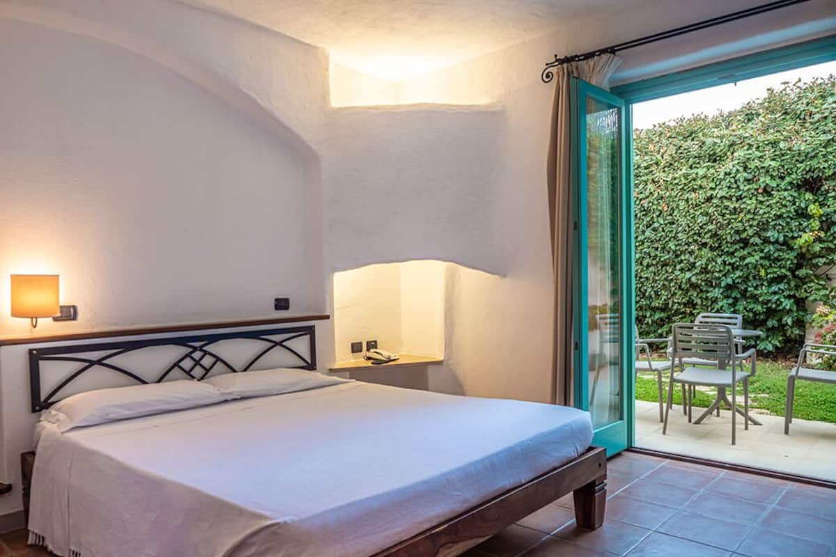 Un dormitorio con cama, lámparas de noche y vista a un patio con mobiliario exterior.