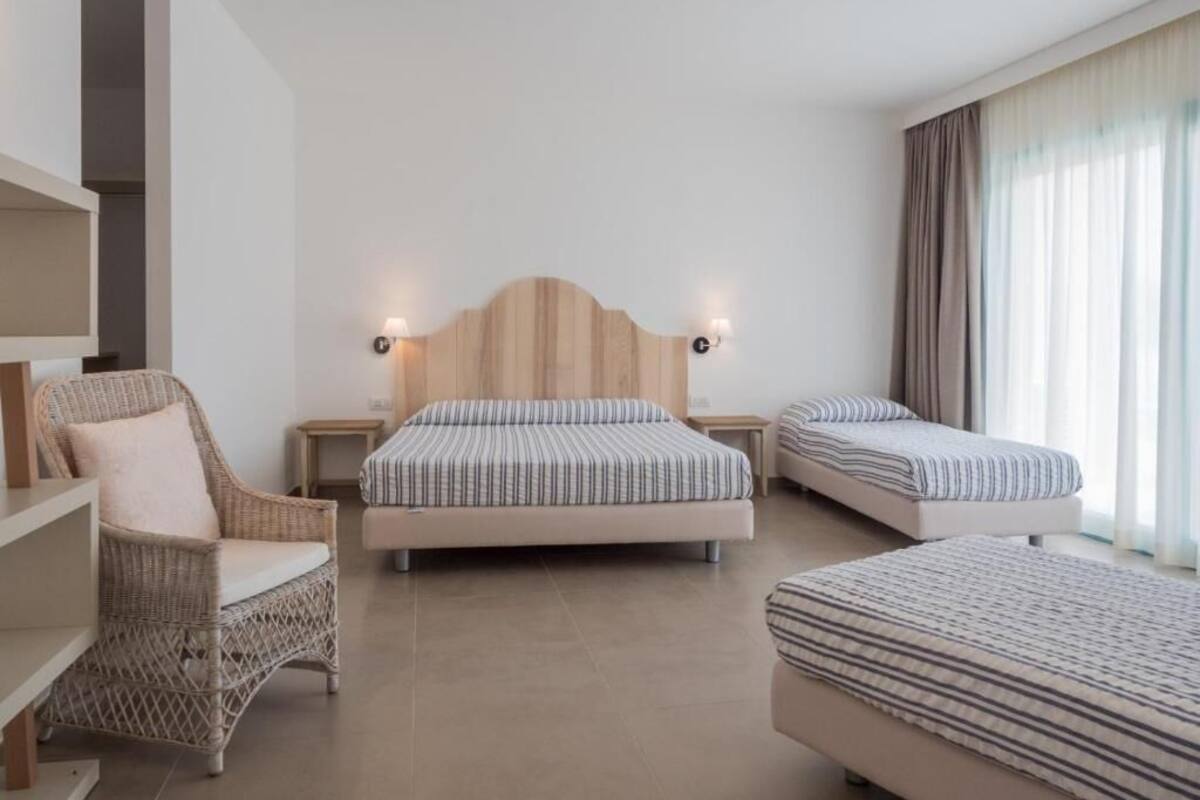 Habitación de hotel con una cama grande, dos camas más pequeñas, una silla de mimbre, una mesita de noche y un gran ventanal con cortinas.