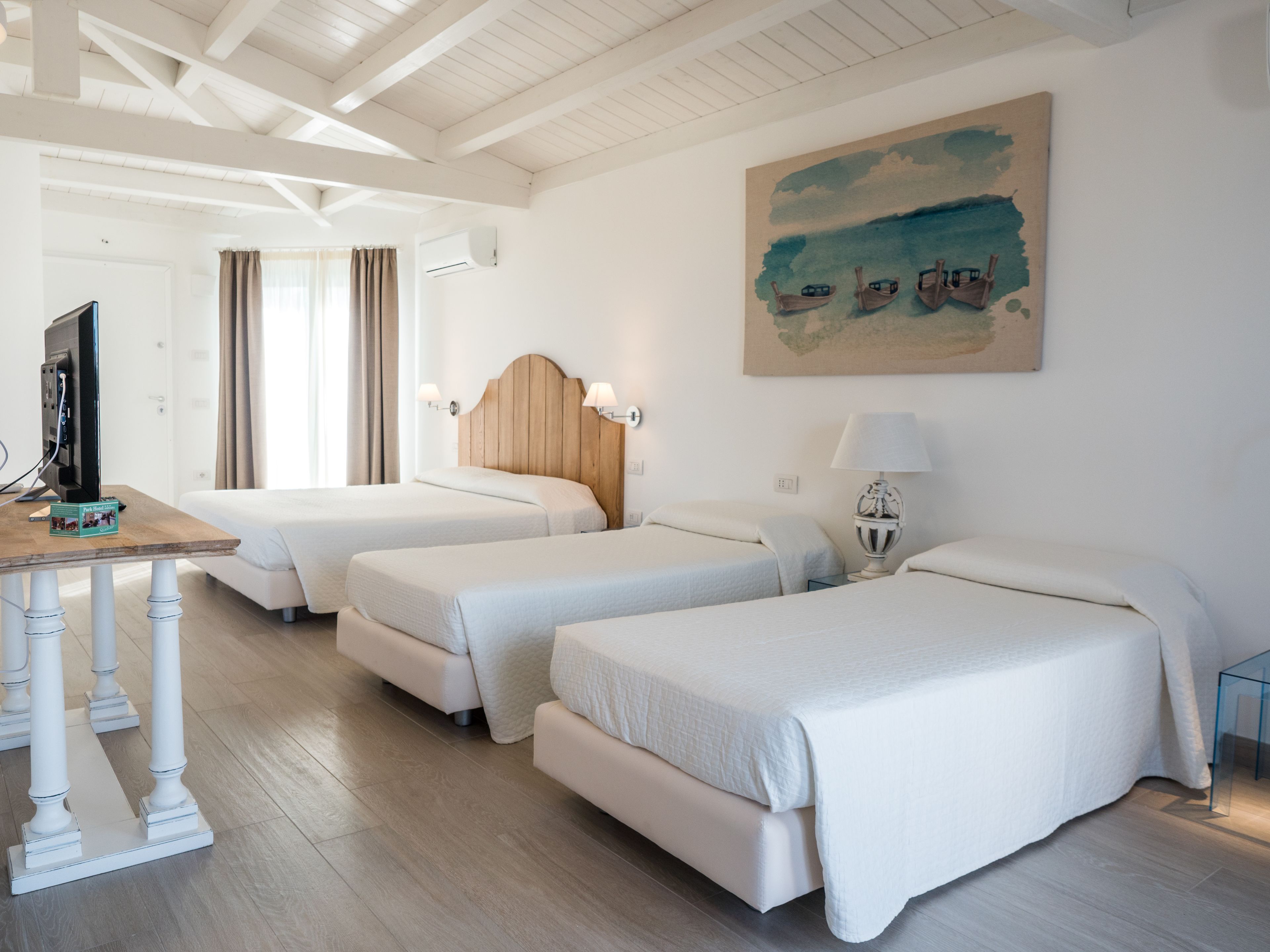 A hotel room with three beds, a desk with a computer, a painting on the wall, and a window with curtains.
