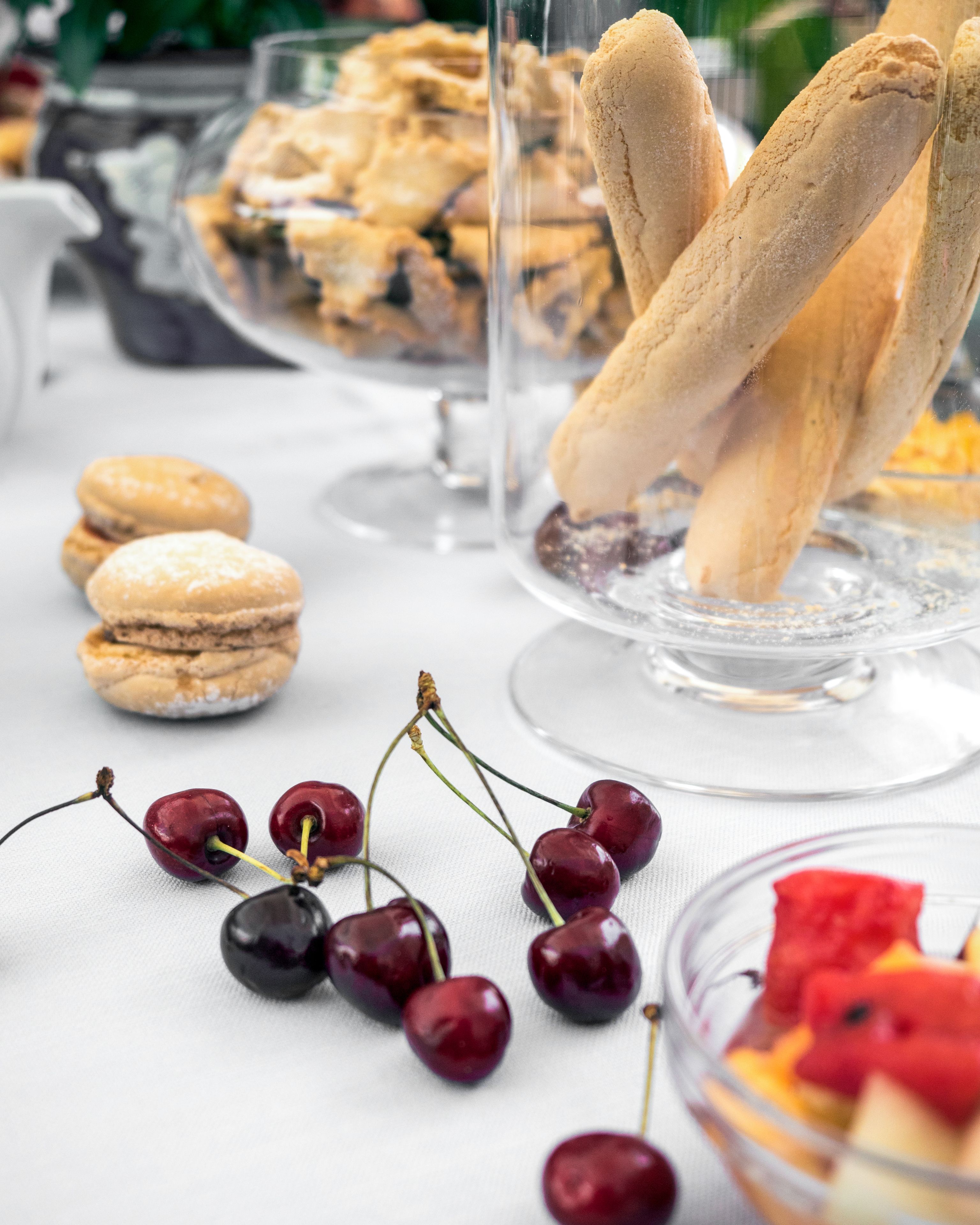 A table with various desserts including macarons, cherries, and a glass dish with breadsticks.
