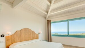Un dormitorio con cabecera de madera, una cama con colcha blanca, una mesita de noche con lámpara, un ventanal con vista al mar y un techo con vigas de madera.