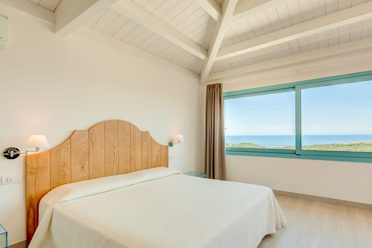 Un dormitorio con cabecera de madera, una cama con colcha blanca, una mesita de noche con lámpara, un ventanal con vista al mar y un techo con vigas de madera.
