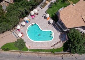 Vista aérea de una piscina rodeada de sillas de descanso y sombrillas, con un edificio al fondo.