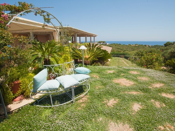 Un jardín con un banco de metal y una pérgola, rodeado de vegetación exuberante y una vista despejada del mar.