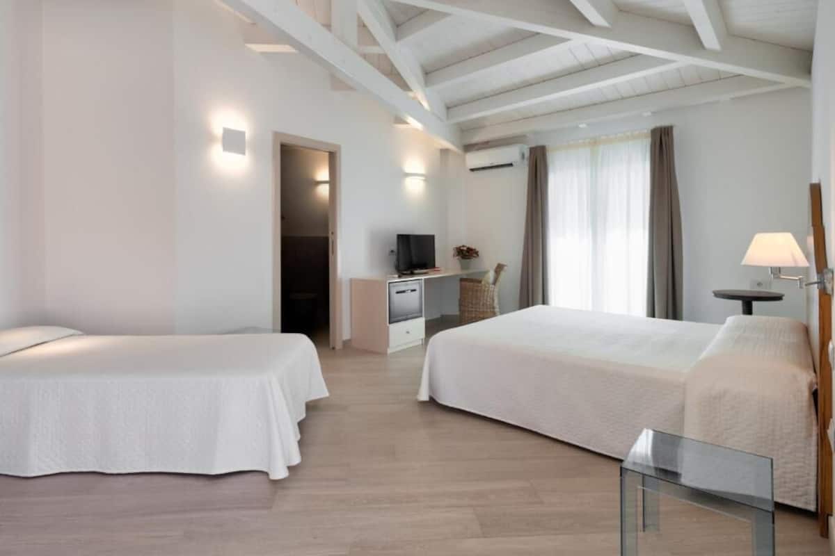 Habitación de hotel con dos camas, un escritorio con televisor, una lámpara y un ventanal con cortinas.