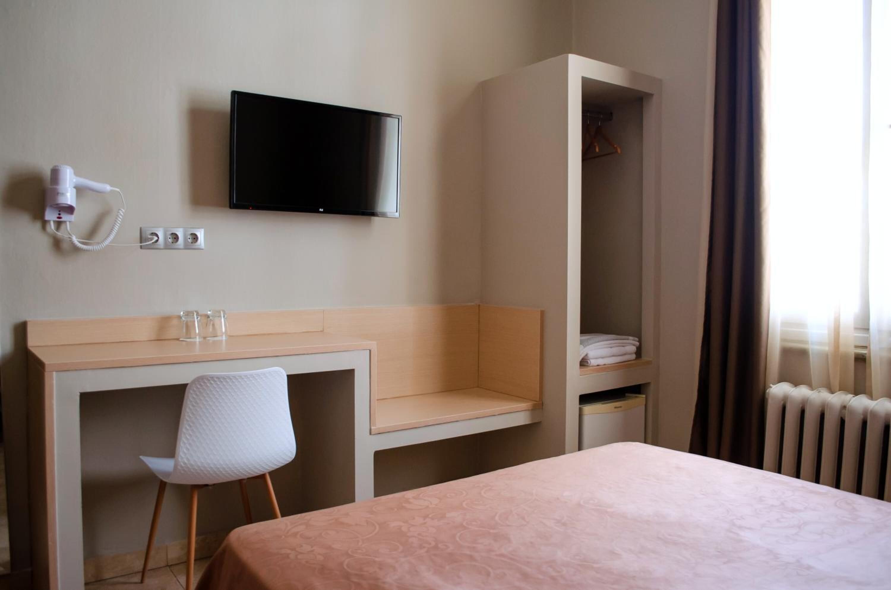A hotel room with a bed, a desk with a chair, a television, a coat rack, and a radiator.