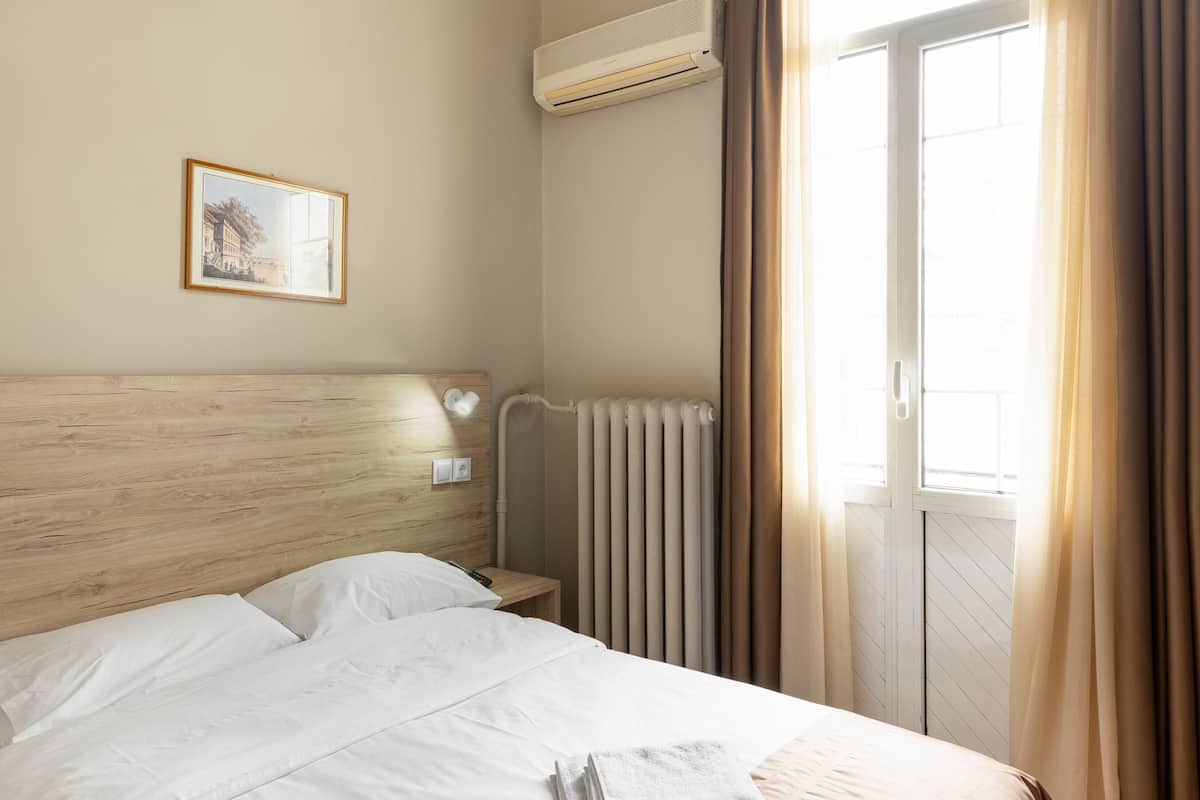 Una habitación de hotel con cama, radiador, equipo de aire acondicionado, ventana con cortinas y un cuadro en la pared.