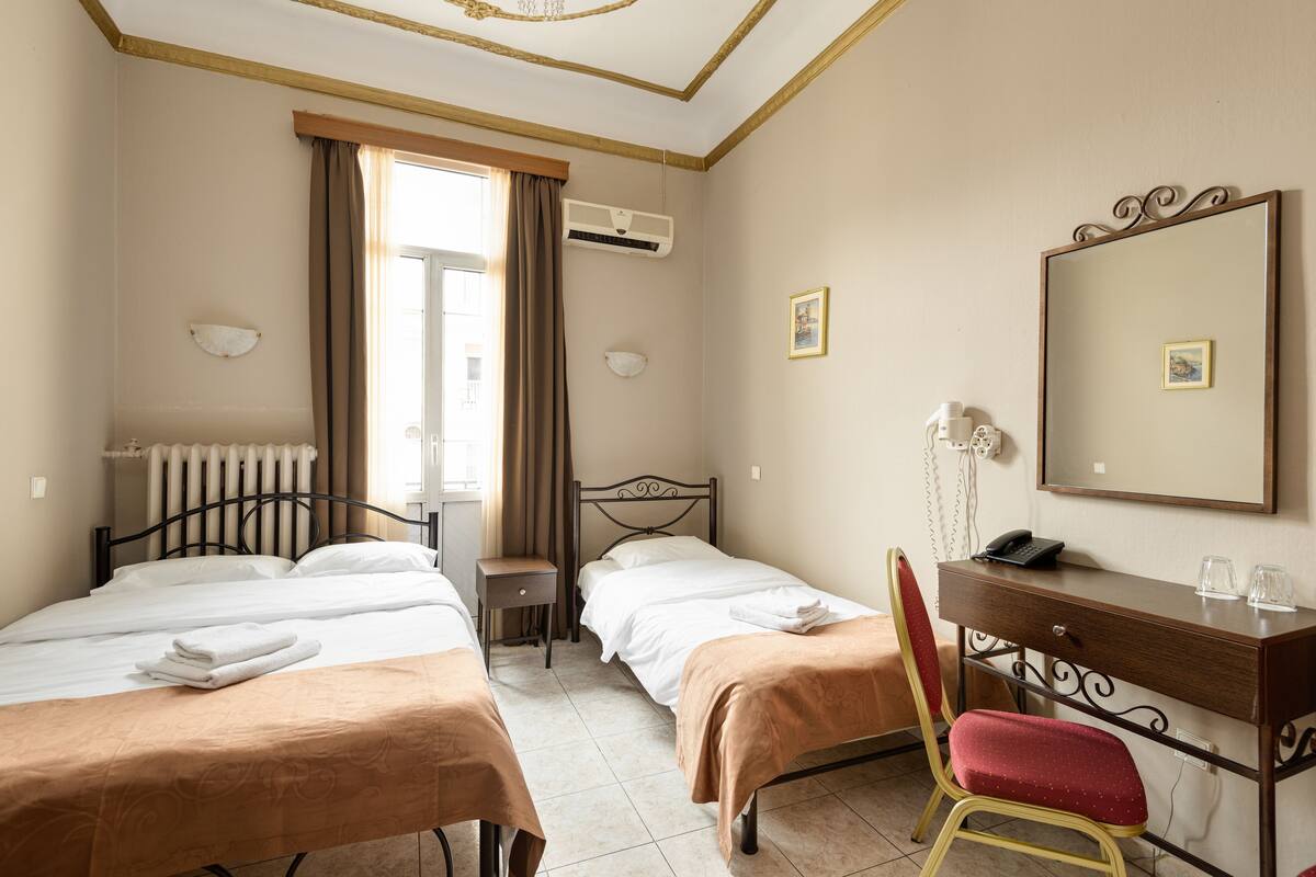Habitación de hotel con dos camas, un escritorio, una silla, un aire acondicionado, un radiador y una ventana con cortinas.