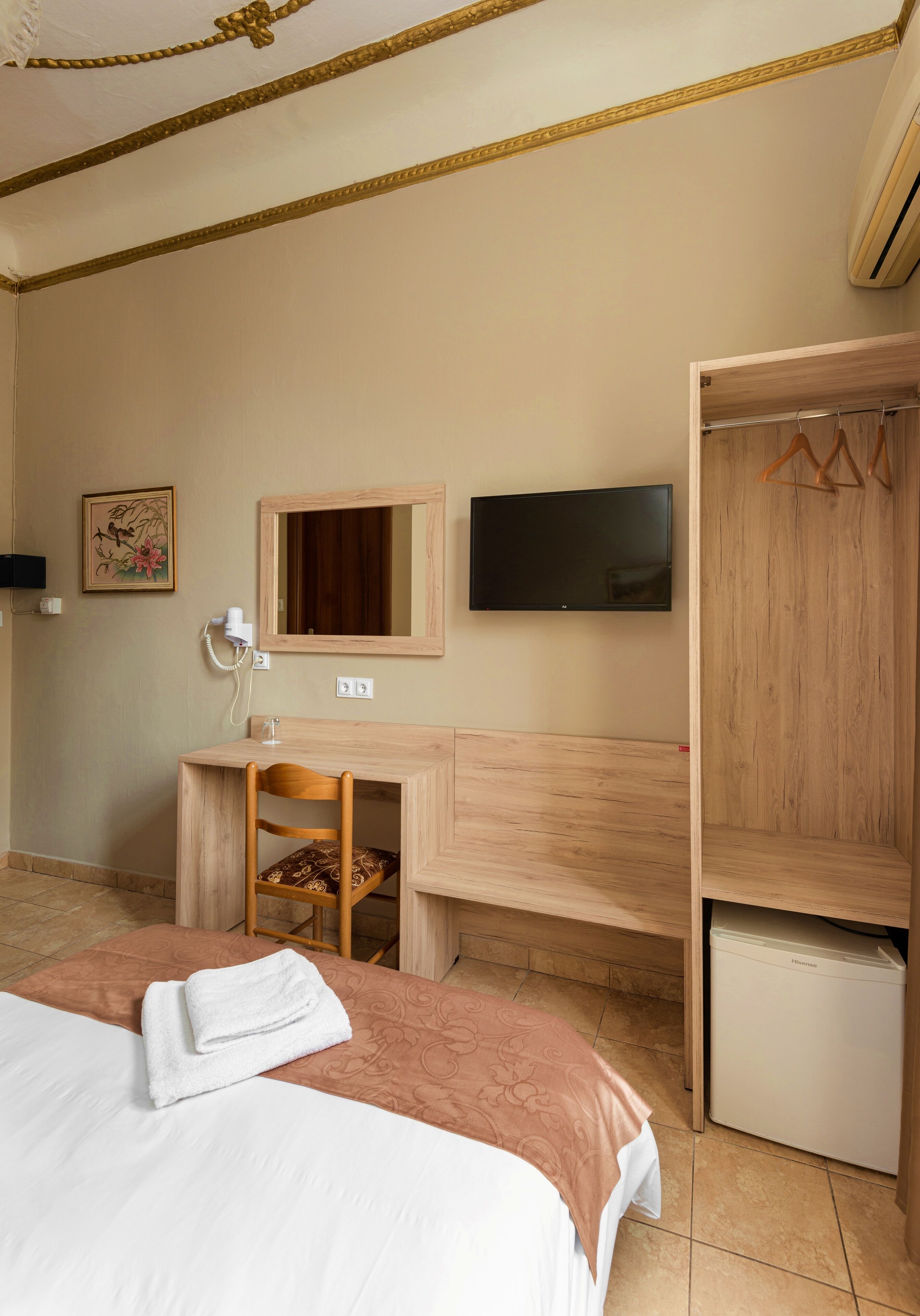 A hotel room with a bed, a desk with a chair, a television, a wardrobe, and a small refrigerator.