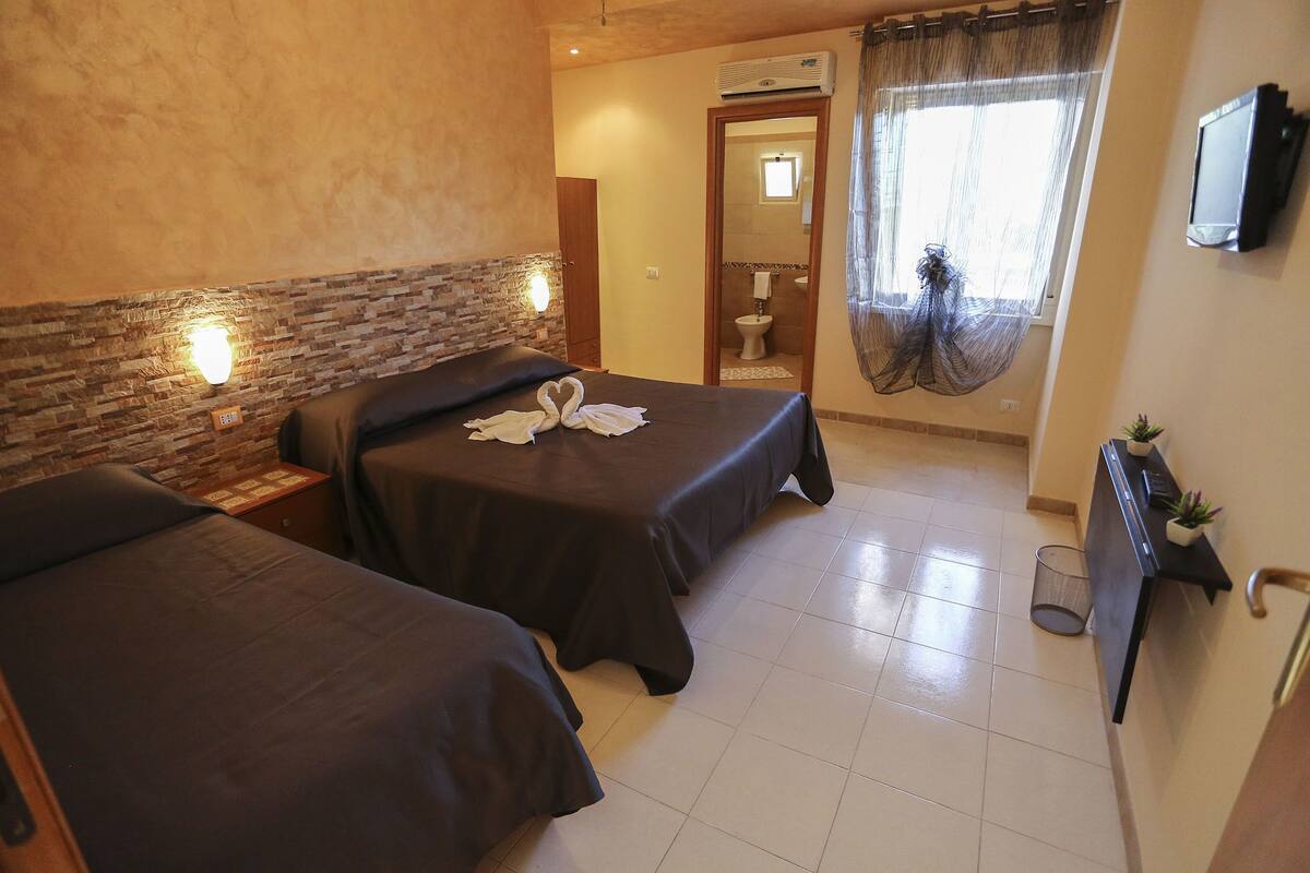 A hotel room with two beds, a TV, a small plant, and a bathroom visible through an open door.