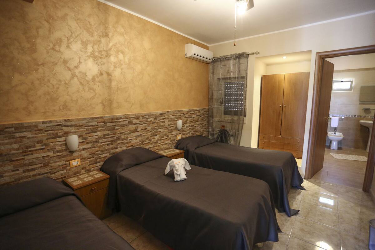 A hotel room with two beds, a stone accent wall, an air conditioning unit, a wooden wardrobe, and a bathroom visible through an open door.