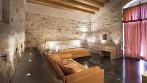 Un dormitorio con pared de piedra, vigas de madera en el techo, una cama, una mesita de noche y una ventana con cortinas.