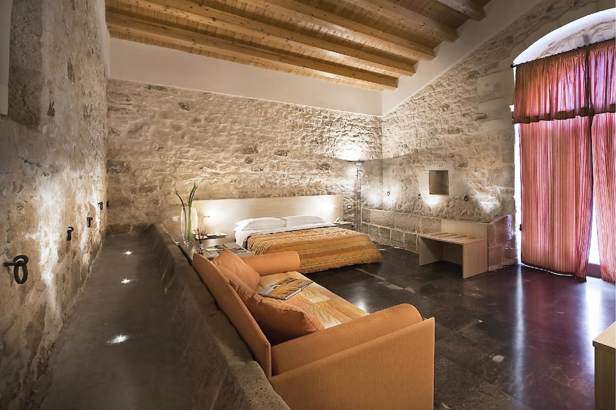 Un dormitorio con pared de piedra, vigas de madera en el techo, una cama, una mesita de noche y una ventana con cortinas.