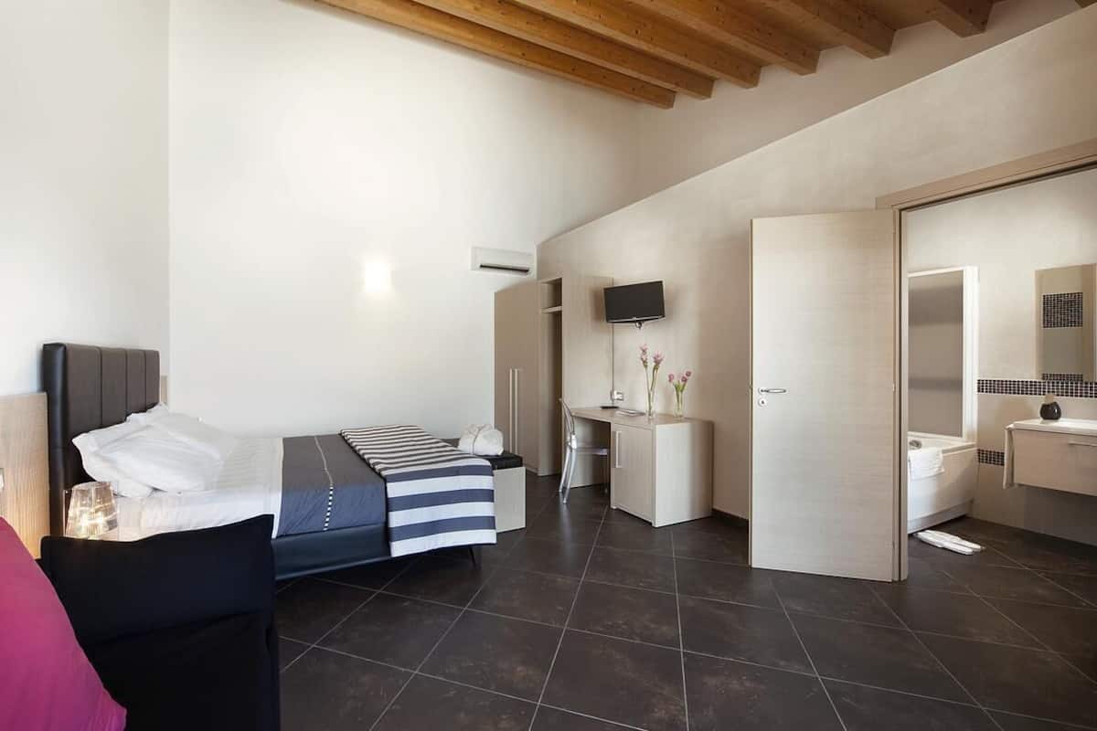 Un dormitorio moderno con cama, mesita de noche y escritorio con silla. Cuenta con un baño con inodoro, lavabo y espejo.