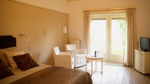 Une chambre avec un lit, deux chaises blanches, une petite table ronde avec une bouteille, un téléviseur posé sur un meuble et une porte-fenêtre coulissante donnant sur un espace extérieur.