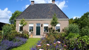Une maison à deux étages, construite en briques, avec un toit recouvert de tuiles sombres, une porte verte et un jardin devant la maison où poussent des rosiers et de la lavande.