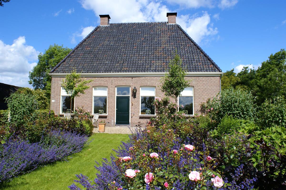 A two-story brick house with a dark tiled roof, a green door, and a front garden with roses and lavender.