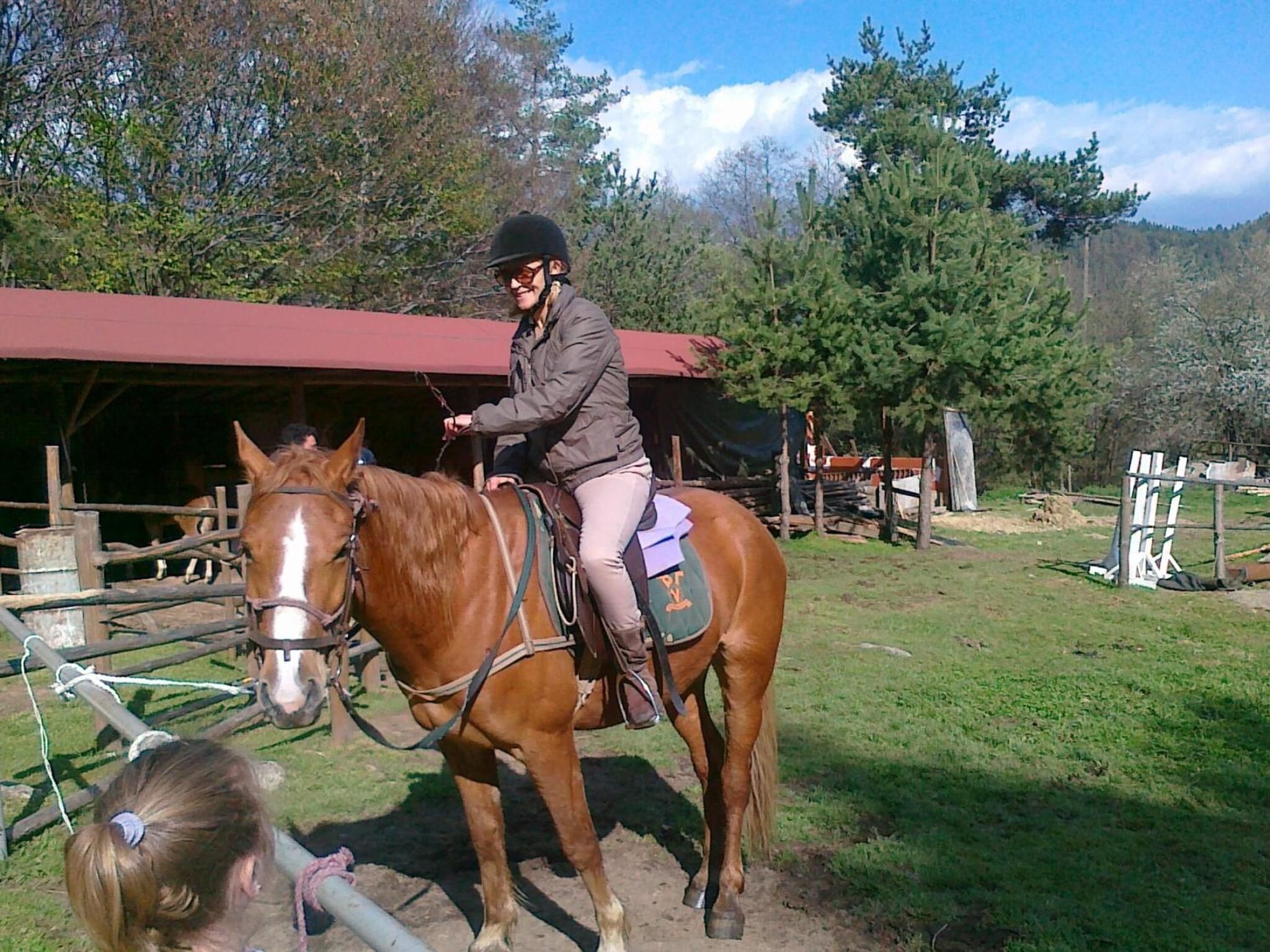 Une personne à cheval, évoluant dans un environnement extérieur, avec une écurie et des arbres en arrière-plan.