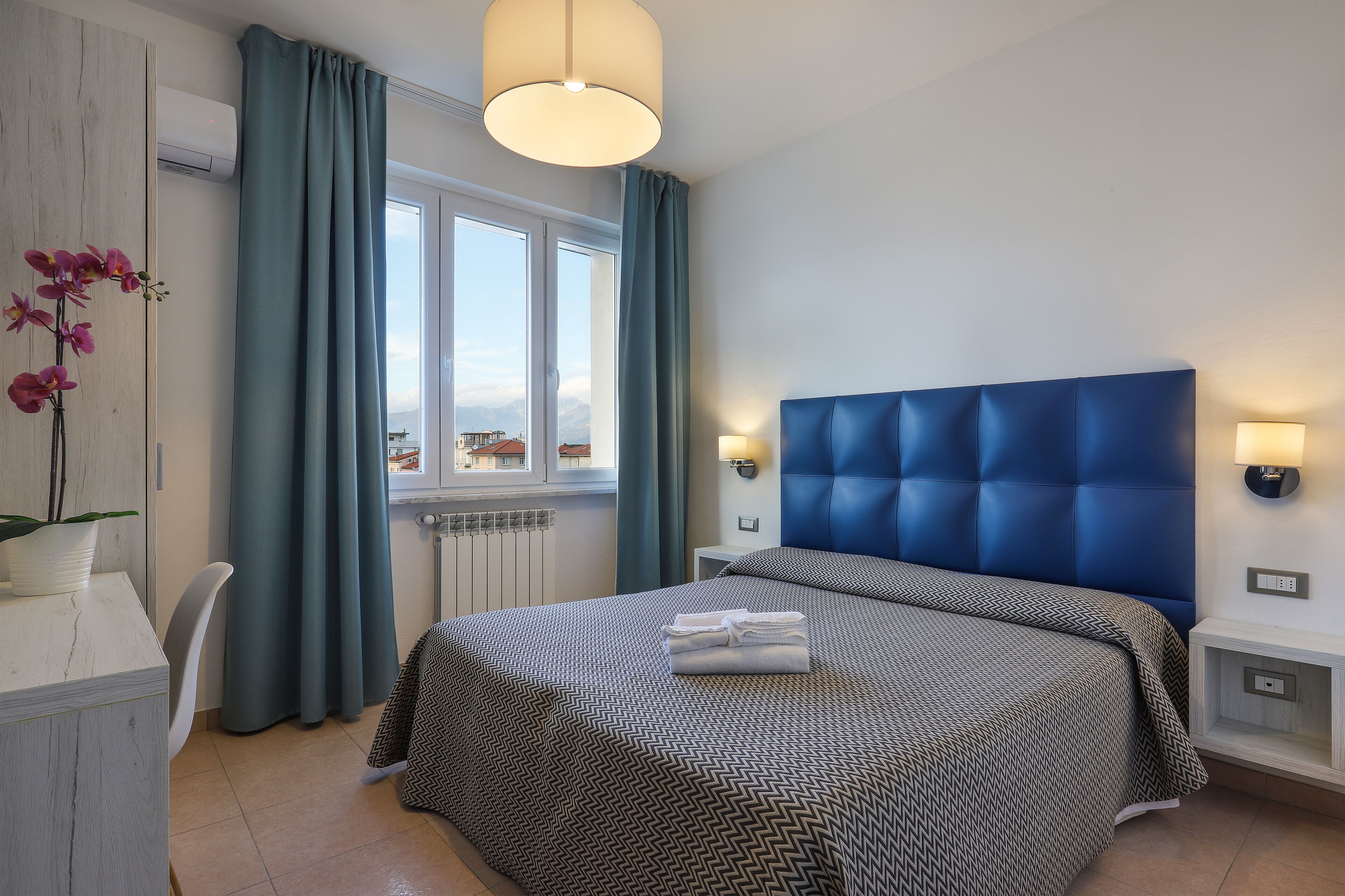 Habitación de hotel con una cama grande, un escritorio con silla, una ventana con cortinas y vistas a edificios.