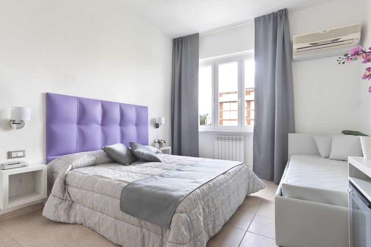 A modern bedroom with a large bed, a bedside table, a desk, and a window with curtains.