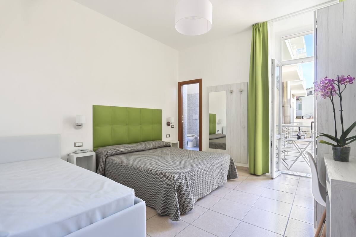 A hotel room with a bed, a bedside table, a mirror, a green curtain, and a potted plant.