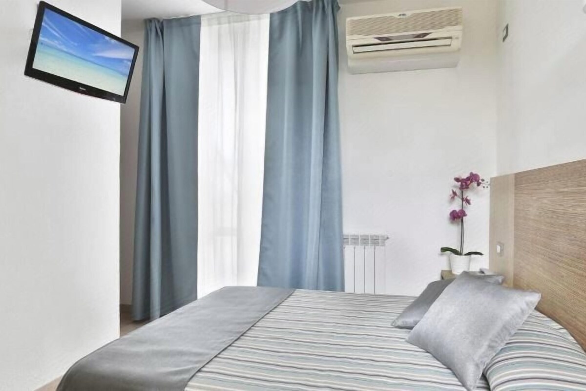 Habitación de hotel con cama, cortinas, televisor montado, equipo de aire acondicionado y una orquídea en maceta.