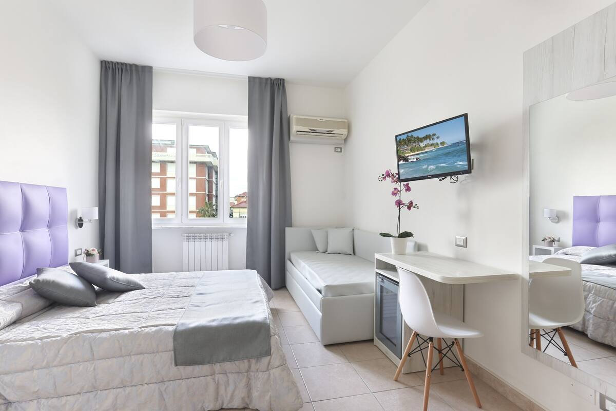 A modern hotel room with a bed, a desk, a chair, a TV, and a window with curtains.