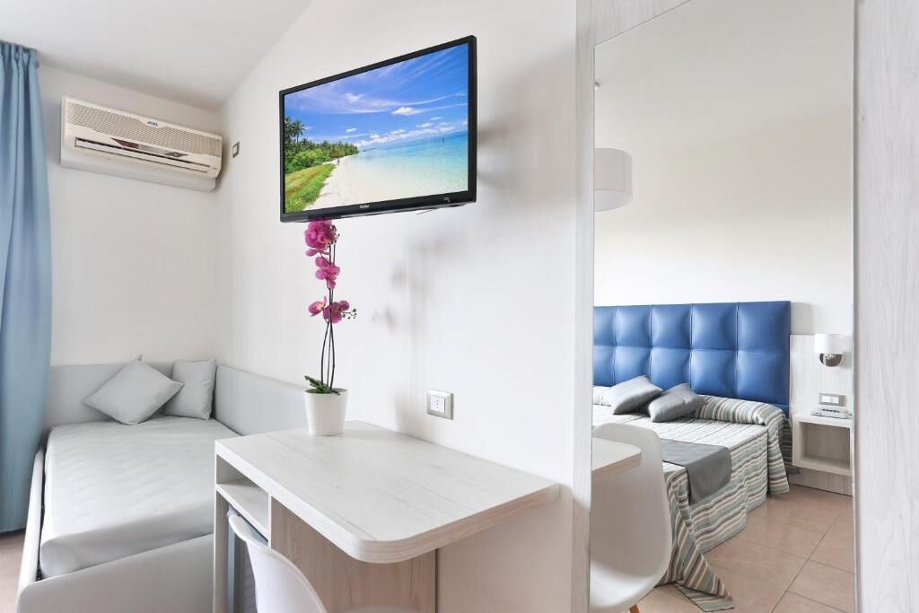 A hotel room with a bed, a desk, a chair, a TV, and an air conditioner.